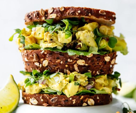 One chicken salad sandwich, made on whole grain bread, stacked on top of another with a white background. 