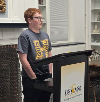 Ryan Schlosser at a podium in the Community Room, reading aloud his Grant Sweet essay entry. 