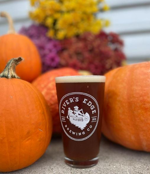 A pumpkin ale in a River's Edge pint glass, situated on the REBC deck with mums and pumpkins behind it. 