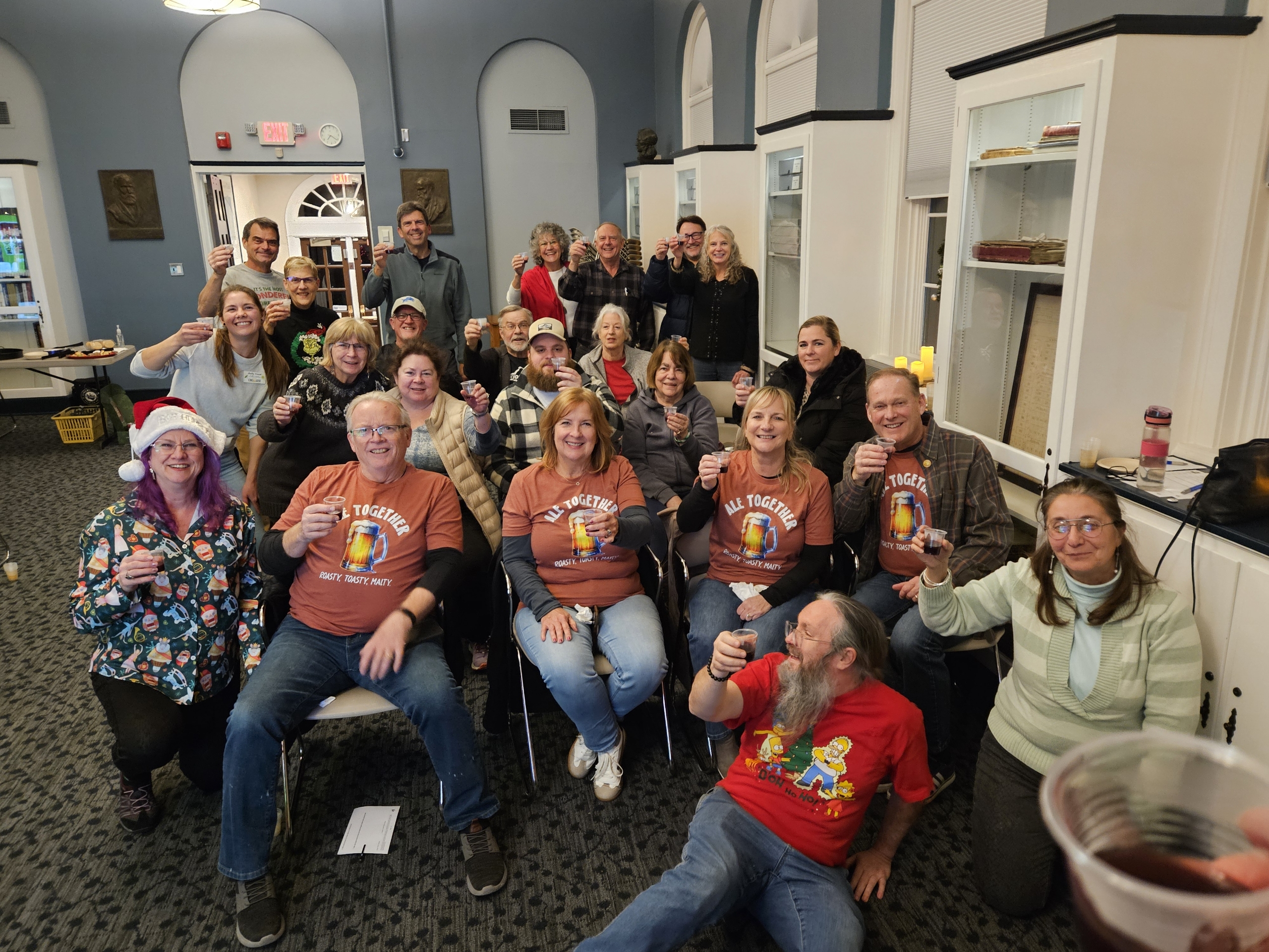 The Ale Together Now crew in the Community Room, toasting the camera with a small cup of wassail. 