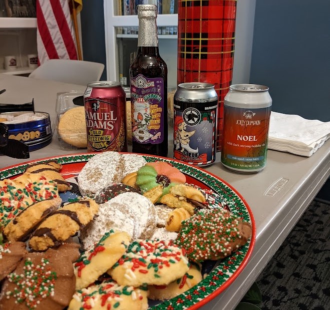 Holiday cookies on a platter in front of four Christmas ale cans and a red plaid thermos, on a table in the Community Room. 