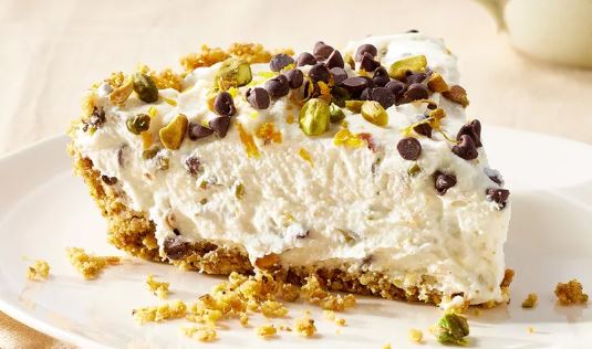 A slice of cannoli cream pie on a white plate, topped with mini chocolate chips and pistachios. 