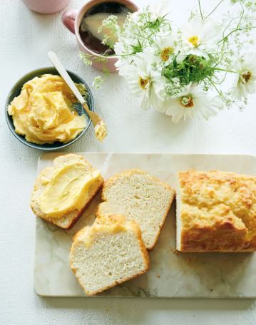 A biscuit loaf, partially sliced on a marble cutting board, with a small dish of honey butter, a mug of coffee, and a small vase of white cosmos. 