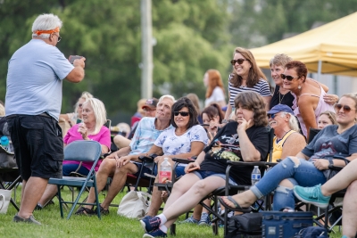 A man taking a picture of a group of friends, seated in lawn chairs and standing behind, on the South Lawn of the Library during a Summer Concert.