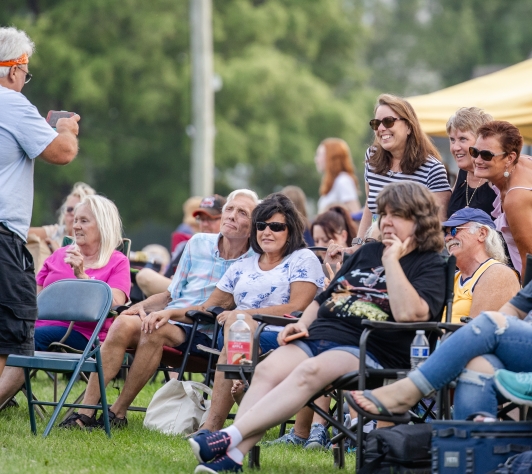 A man taking a picture of a group of friends, seated in lawn chairs and standing behind, on the South Lawn of the Library during a Summer Concert. 