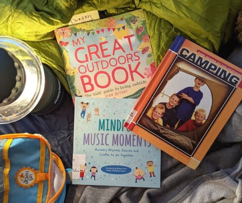 Camping books in a pile with a backpack, reading lamp, and sleeping bag. 