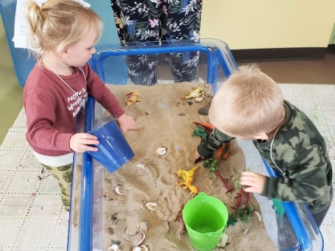 Two children playing in a sensory bin filled with sand and small toys. 