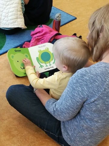 Looking over the shoulder of a women with a baby on her lap, flipping through a frog-themed board book. 