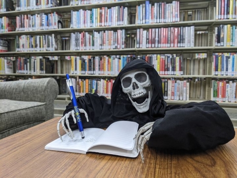 A plastic skeleton holding a Cromaine pen, writing in a notebook in front of the Cromaine bookshelves. 