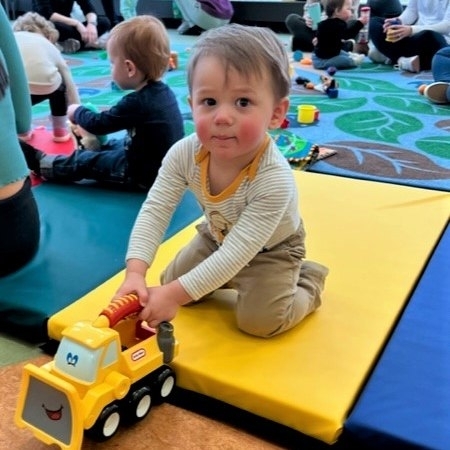 Baby playing with a toy truck on a yellow mat, in Cromaine's Youth Program Room. 