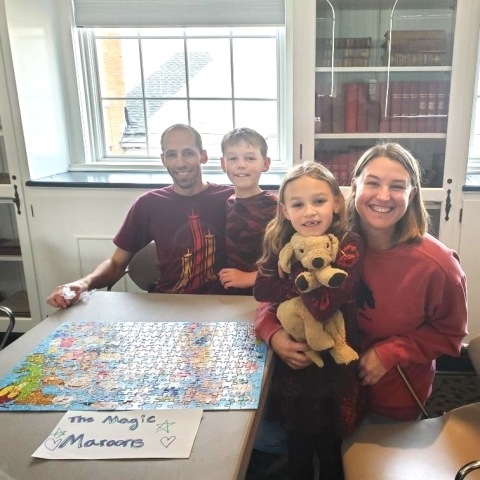 The Magic Maroons, a family of four wearing maroon shirts, sitting at a table around their recently completed puzzle in the Community Room. 