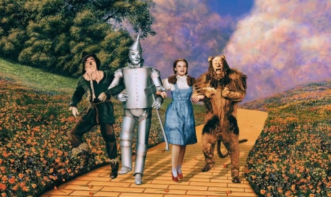 A still of The Wizard of Oz film showing the Scarecrow, Tin Man, Dorothy, and the Lion on the Yellow Brick Road. 