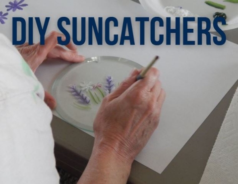 A hand holding a suncatcher in place, while the other hand paints purple flowers onto the suncatcher. 