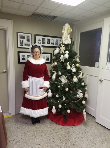 Mrs. Claus standing and smiling next to a decorated Holiday tree. 