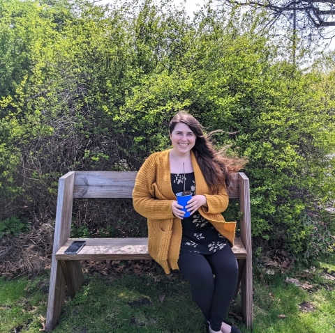 Librarian Heather sitting on a bench in the Library garden, holding a baby tree. 