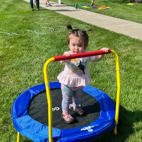 A toddler on a small trampoline, holding onto a rail, on the South Lawn. 