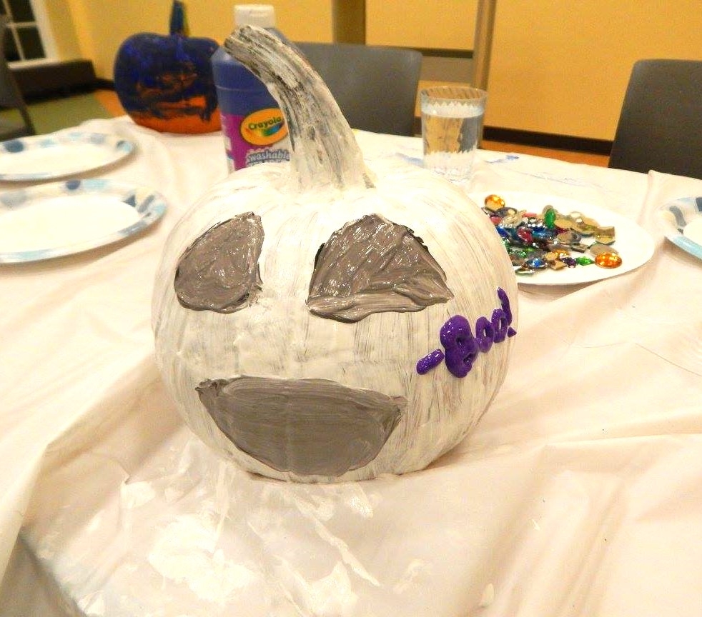 A painted pumpkin, white with silver jack 'o lantern design, and the word 'Boo' in purple, in the Youth Program Room of the Library. 