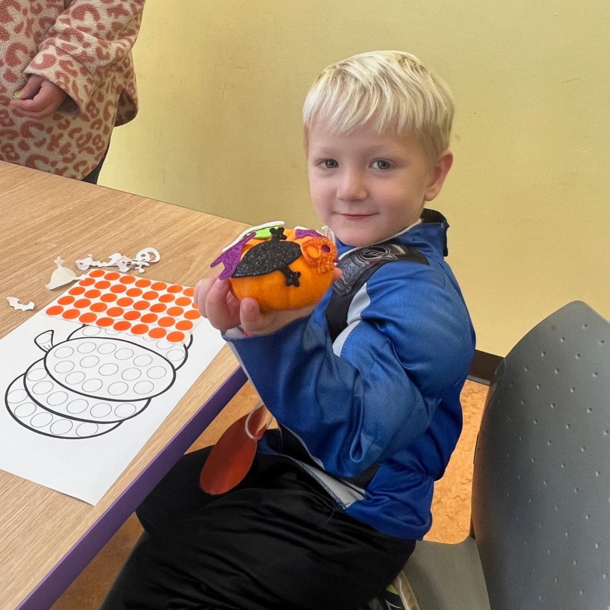 A boy in costume in the Youth Program Room, holding up a pumpkin craft, at a table with Halloween-themed activity sheets. 