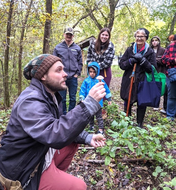 Andrew Fleszar of MI Mushroom Hunters kneeling in the woods and holding up a mushroom, speaking to a group of mushroom hunt attendees. 