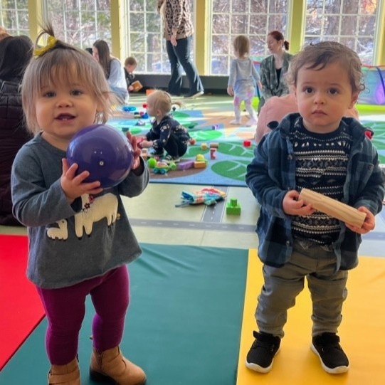 Two toddlers holding up toys in the Youth Program Room of the Library. 