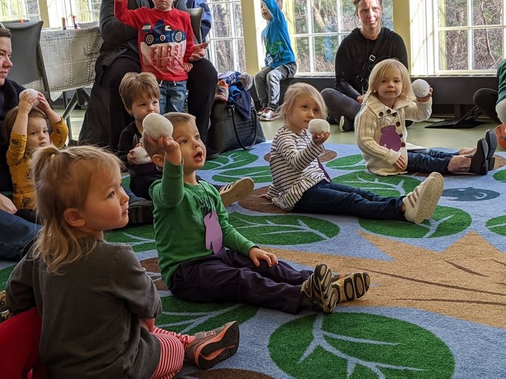 Children sitting on the storytime carpet in the Youth Program Room, holding up wool snowballs. 